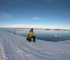 Man in AAD cold weather grea (yellow and black) and climing equipment (helmet, ice axe and crampons on boots) kneels on snow covered slope. Behind is bay of open water with ice cliffs along edge and blue sky above