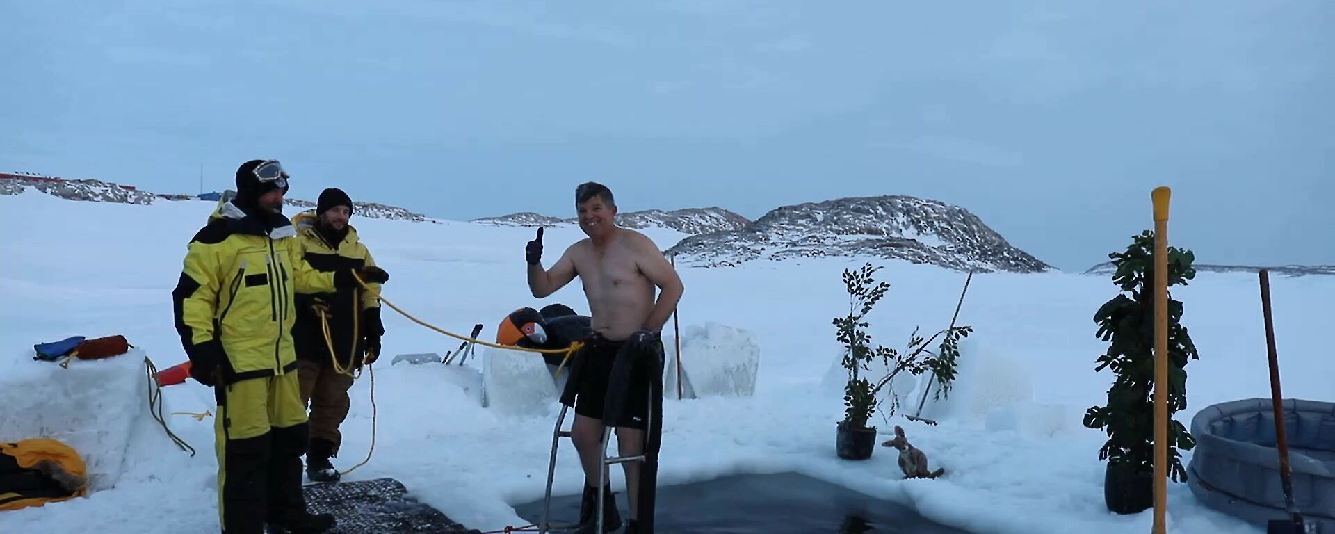 Man in board shorts stands on ladder on edge of hole in the sea-ice. He smiles at camera giving thumbs up. He’s attached by a rope to two safety members, dressed in AAD cold weather jacket and pants (yellow and black)