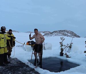 Man in board shorts stands on ladder on edge of hole in the sea-ice. He smiles at camera giving thumbs up. He’s attached by a rope to two safety members, dressed in AAD cold weather jacket and pants (yellow and black)