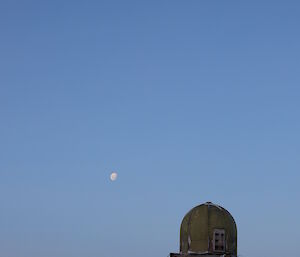 Foreground a green domed building on rocky outcrop. Above clear blue skies with full moon in white centre picture