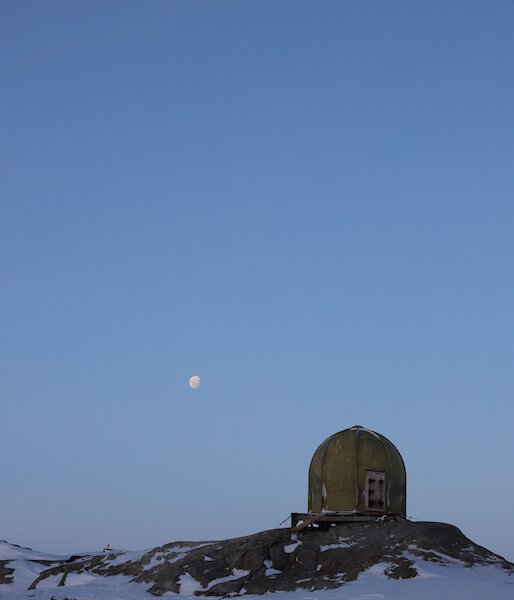 Foreground a green domed building on rocky outcrop. Above clear blue skies with full moon in white centre picture