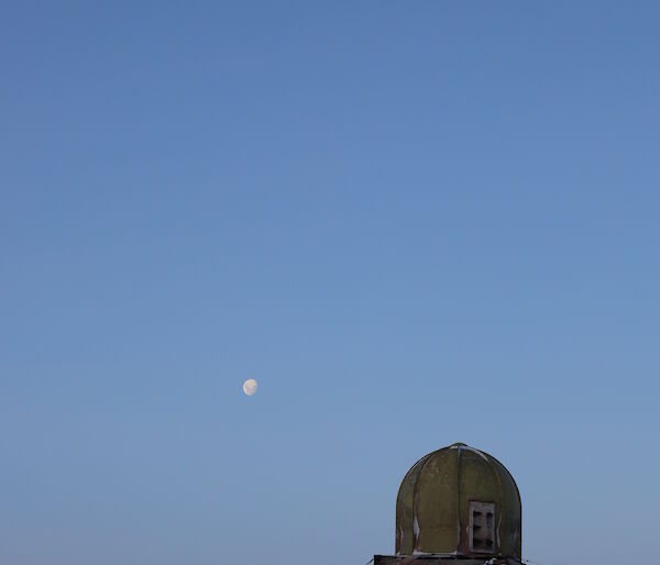 Foreground a green domed building on rocky outcrop. Above clear blue skies with full moon in white centre picture