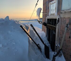 Side of old building with large pile of snow covering over half the door. Shovel tossing scoop of snow out the door