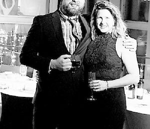 Couple dressed for a formal dnner, man in suit and woman in long halter neck dress, stand together with table behind and fiary lights above