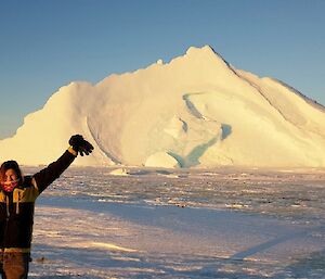 Woman in left foreground raising hand above head, with stranded ice berg behind caught in sea ice. Blue sky above