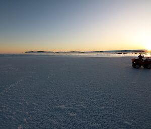 Expanse of flat sea ice extends to horizen where it meets open water and sunset. Clear blue sky above. Sillouetted quad bike and person standing beside taking photo at centre right