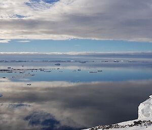 Overlooking mirror like surface of sea with ice covered shore at right. Blue sky with clouds reflected perfectly in the water