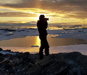 Man in foreground standing on rocky outcrop with camera held up to eye, sillouetted against ice covered water at sunset