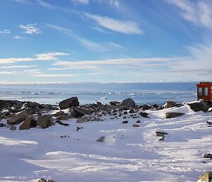 Compartment of a tractor converted into an outhouse sits on rocky outcrop overlooking view of bay with rocky islands, ice bergs and a blue sky with scattered white clouds