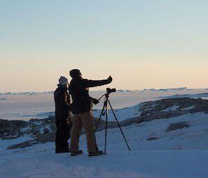 Man and woman stand behind camera on tripod looking over outside scene working out next shot