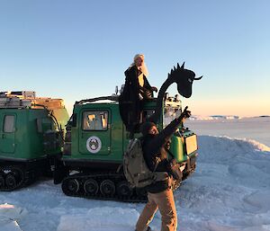 Man straddles roof of hagglunds vehicle with neck and head of dragon attached to front. In front camera man points off into distance