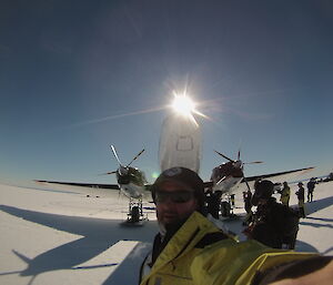 Man in yellow gortex shell jacket and black ball cap standing in foreground taking selfie with arm at full length. In background a basler aircraft front on with white undercarriage on ice runway and with blue sky above