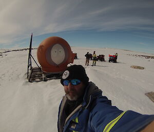 Man in foreground taking selfie at full arm length, in background orange melon hut with face of Kenny from Southpark painted onto the side