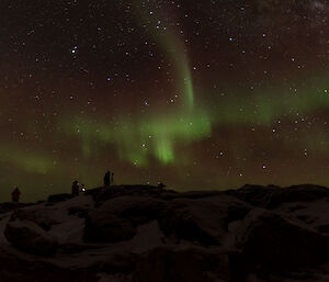 Green and red aurora above a dark horizen that has three people sillouetted standing at camera tripods