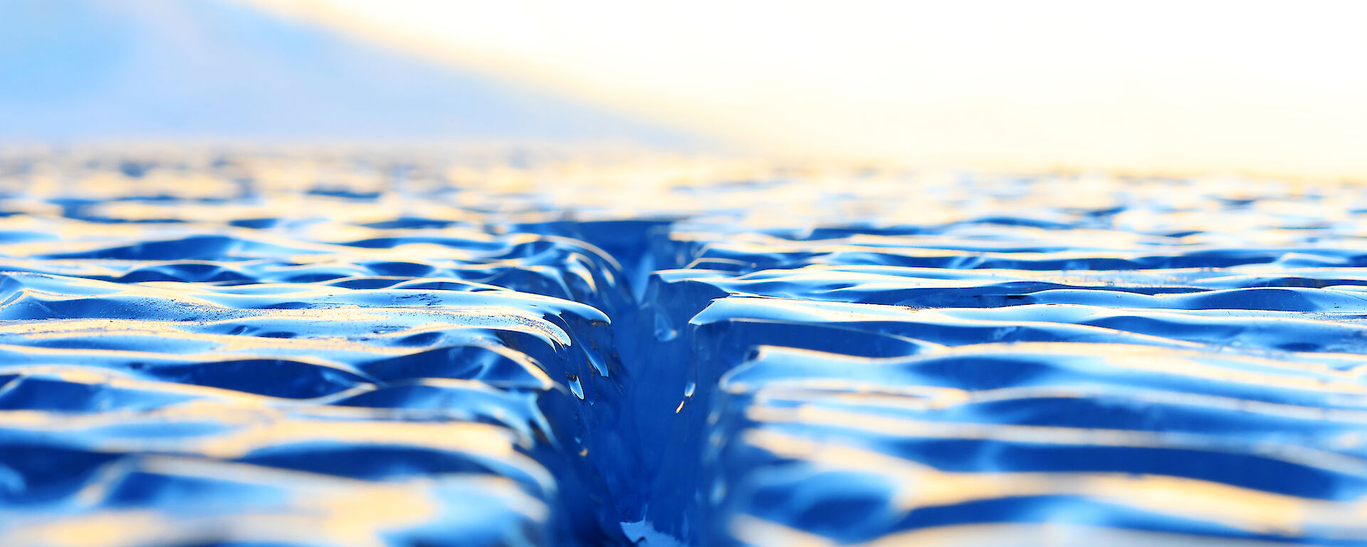Close up view across the surface of the ice lake, showing bright blue smooth undulating surface with a crack down the centre of the frame extending out to the horizen