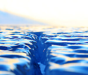 Close up view across the surface of the ice lake, showing bright blue smooth undulating surface with a crack down the centre of the frame extending out to the horizen