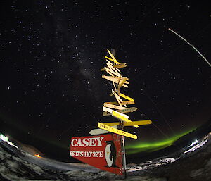 Fish eye shot of Casey sign lit up on foreground with night sky above and bright green aurora on the distant horizen