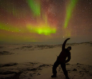 Woman in foreground doing yoga pose (side bend) while above in the night sky is a yellow, green and red aurora