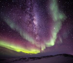 Small amount of snow covered rocky outcrop in foreground, above the expanse of night sky filled with three waves of yellow-green and red aurora surrounding the milky way through centre of picture