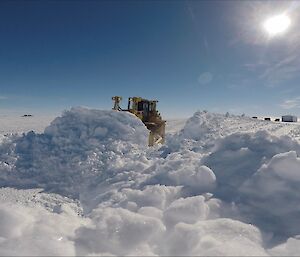 Large mound of broken up snow in foreground, with bulldozer pushing snow towards camera, in the distance the aerodrome camp, above blue skies and sun