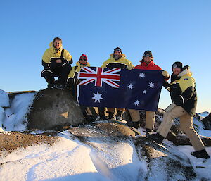 Group of five men standing on rocky outcrop in cold weather gear holding up Australian flag between them