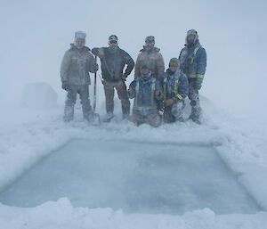Group of men in cold weather gear covered in snow, standing behind a large square hole cut in the sea ice, weather around is foggy with reduced visibility