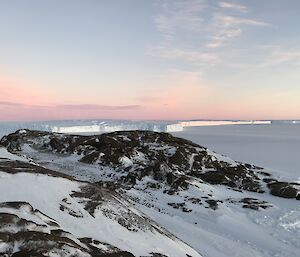 Foreground is large rocky hill with snow, in the distance large bay covered in ice stretching to glacier edge, with pink tinge of sunset above