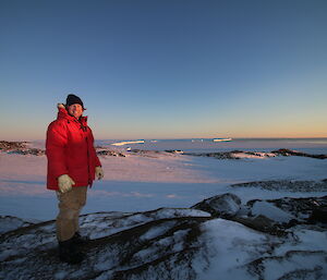 Man standing in foreground with large red down jacket, smiles at camera, in the background snow covered landscape with rocky hills, sea ice and glacier with clear blue sky at twilight with orange tint of sunset