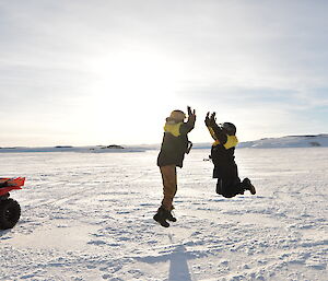 Two expeditioners on sea-ice beside quad bike jumping into air and clapping hands together