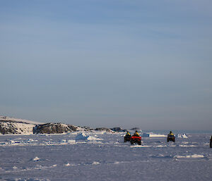 Quad bikes travelling through the sea-ice which has small pieces of ice bergs trapped in the ice, in the distance a rocky peninsula, blue sky above