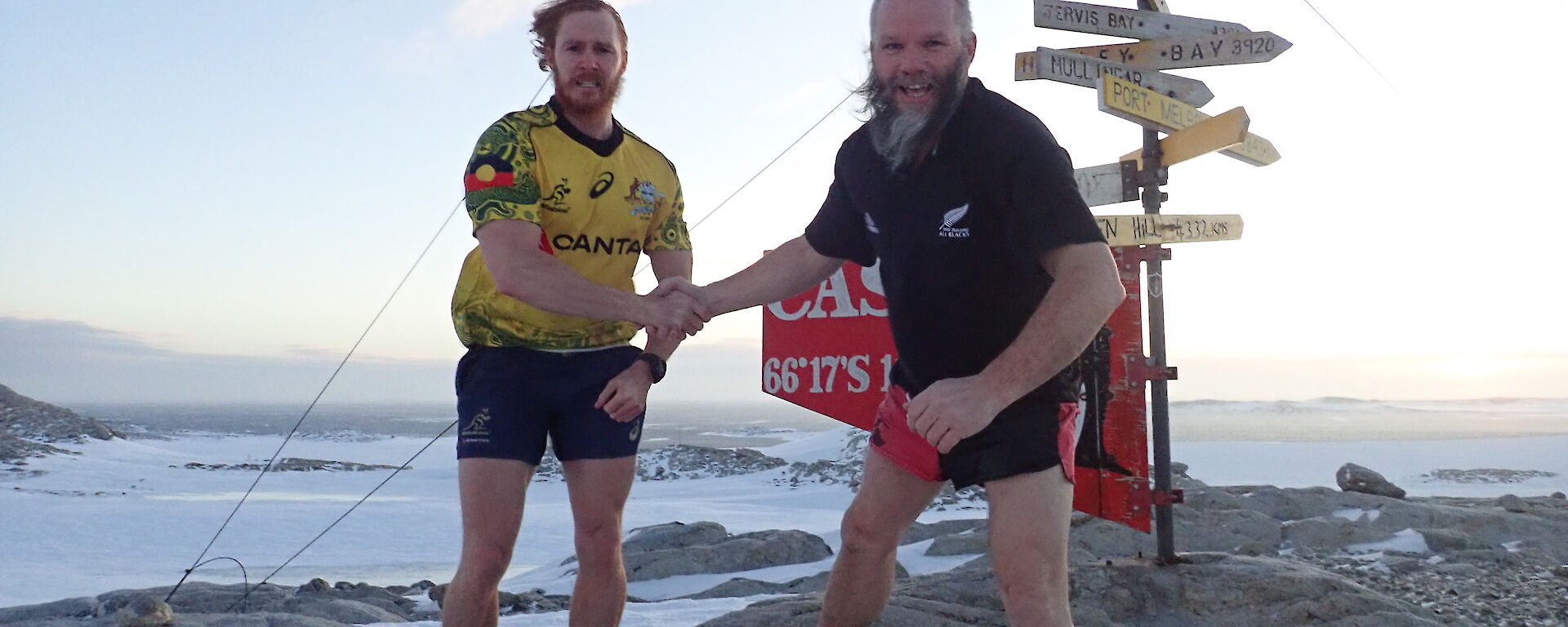 Two men in Wallabies and All Blacks t-shirts and shorts shake hands standing in front of the Casey sign