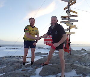 Two men in Wallabies and All Blacks t-shirts and shorts shake hands standing in front of the Casey sign