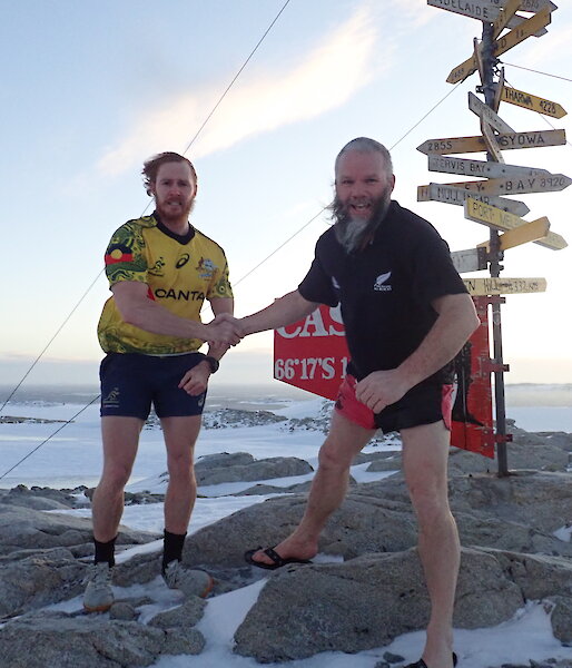 Two men in Wallabies and All Blacks t-shirts and shorts shake hands standing in front of the Casey sign