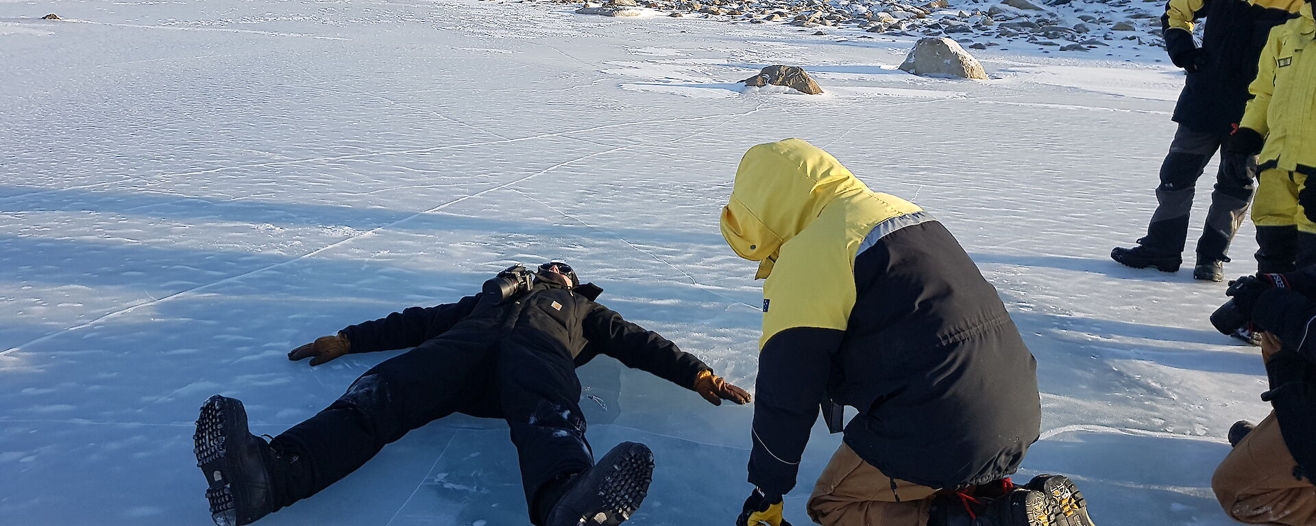 Man lies flat on his back on an ice lake while other expeditioners (3) look on
