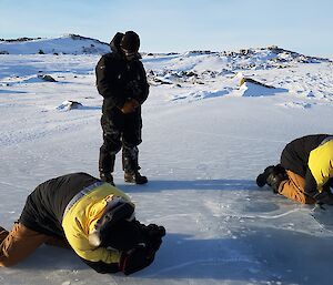 Two expeditioners kneeling down on ice lake trying to take close up photos of the ice, with one man standing looking on