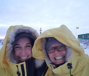 Close up of two female expeditioners with down jackets with fur lined hoods up, smiling at the camera, in the background the Casey sing and flag poles