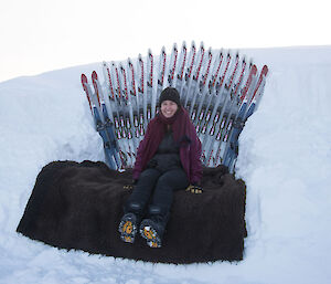 Woman sits in cold weather gear and boots on oversized seat cut into the snow, with skis positioned around the back of the seat to make it look like the Iron Throne from Game of Thrones