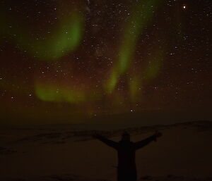 Woman in black sillouted under a night sky with pale green and red aurora above