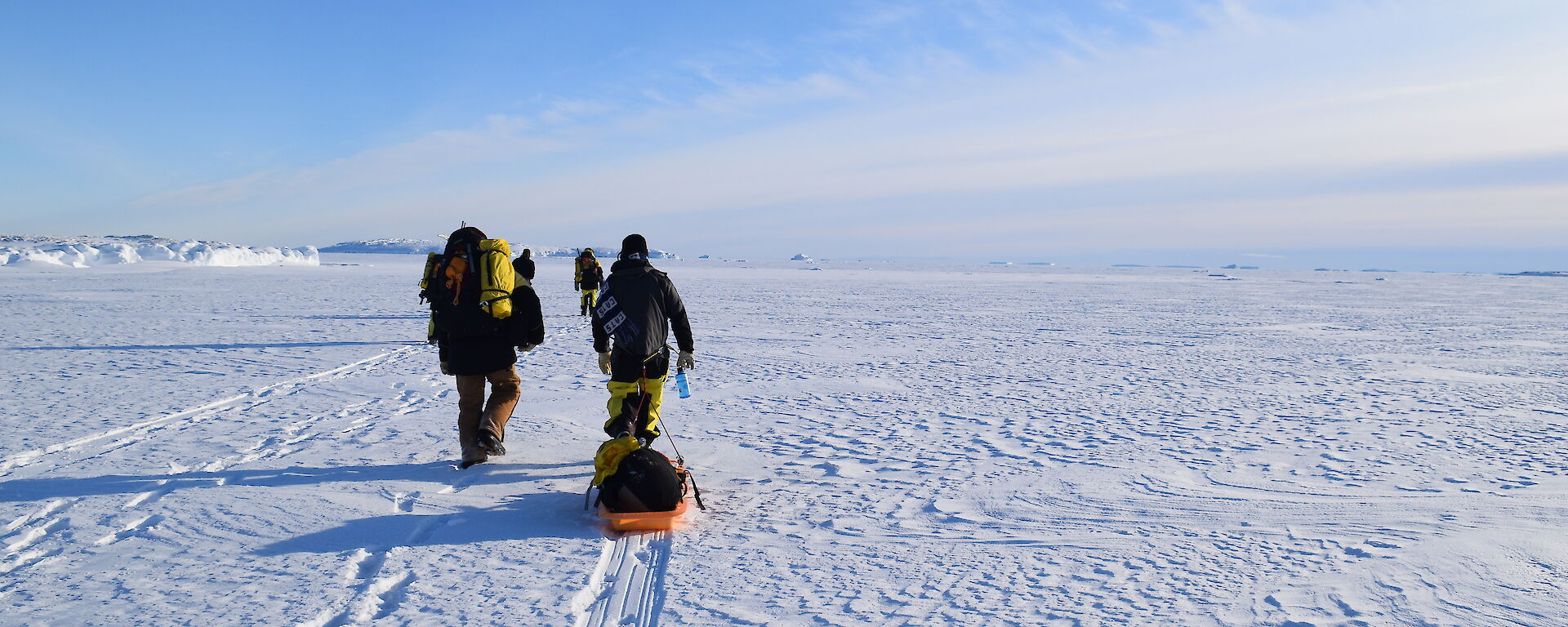 Flat expanse of sea-ice with people walking across carrying pack and pullijng sled