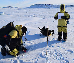 Man on knees measures thickness of sea-ice through a drilled hole in the ice, a second man stands and records measurement onto a piece of paper with his pack beside him on the ice