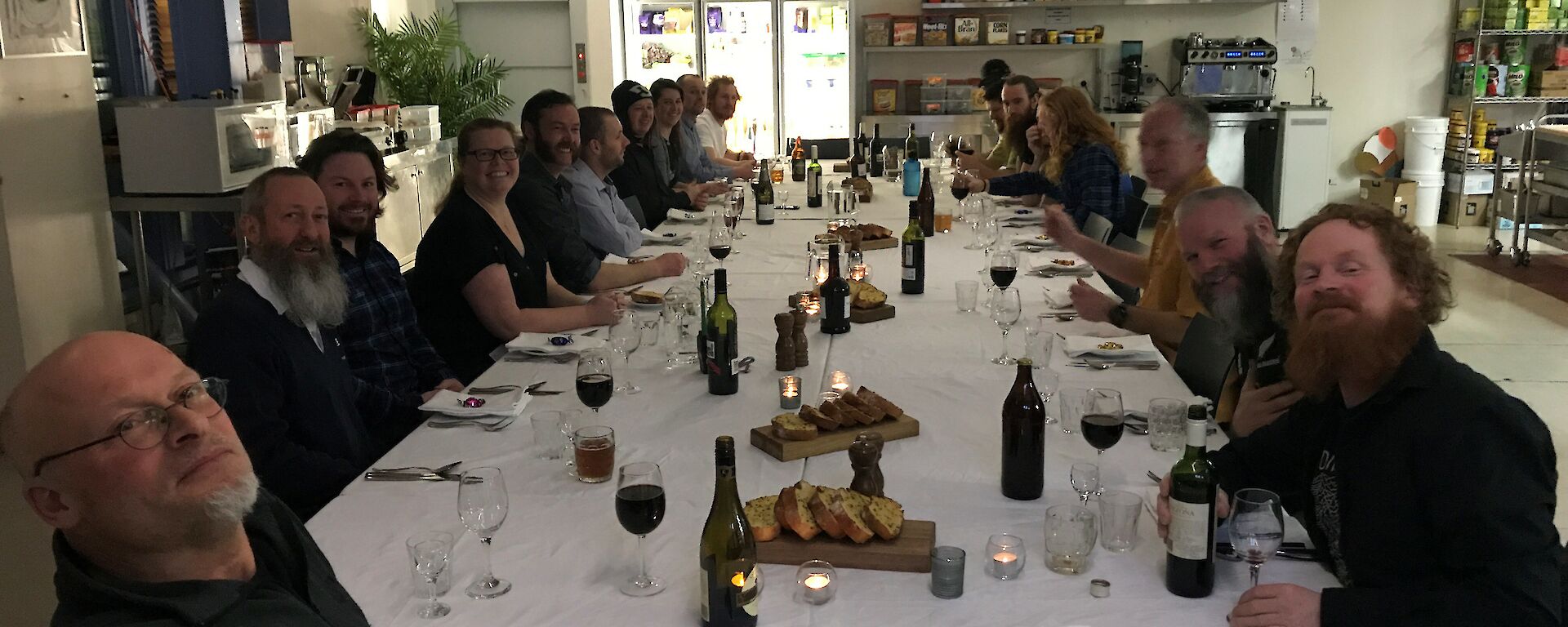 A long table sitting over twenty, set with white tablecloth and crockery, cutlery, napkins, glasses. All twenty people sitting around looking at camera