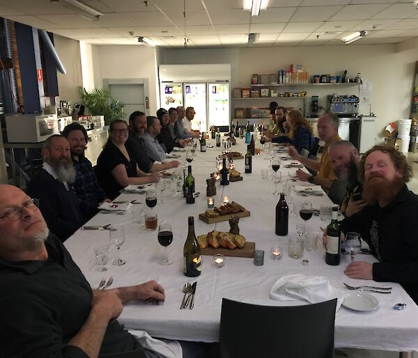 A long table sitting over twenty, set with white tablecloth and crockery, cutlery, napkins, glasses. All twenty people sitting around looking at camera