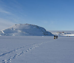 Expanse of flat sea-ice leading to snow covered hill and in the distance the station can be seen, blue skies above. Two sets of tracks leading from front left of picture to centre right where two people are see in the distance