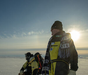 Close up of two men looking off into the distance, while sea-ice extends to horizen and sun sillouettes them
