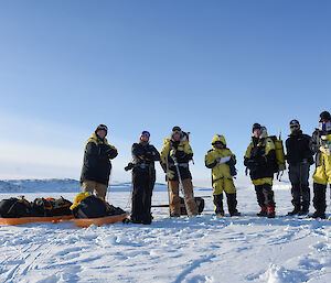 A group of expeditioners standing in a line facing camera with sleds pulled in front. On sea ice with blue sky above.
