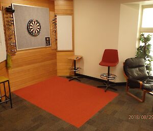 Corner of bar area showing wood panneling behind darts board and score boards, red square of carpet depicting playing area and grey carpet around, scattering of seating