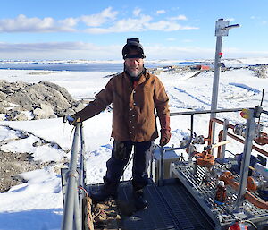 Man standing in boilermaker protective equipment — leather overjacket, welding mask and gloves — on platform over the top of the upper fuel farm with newcomb bay in the background