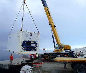 Large yellow crane at edge of warf lifting white shipping container onto barge