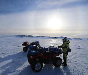 Foreground, two quad bikes with one man standing beside, on expanse of sea-ice, in the distance the sun above the horizen with halo around