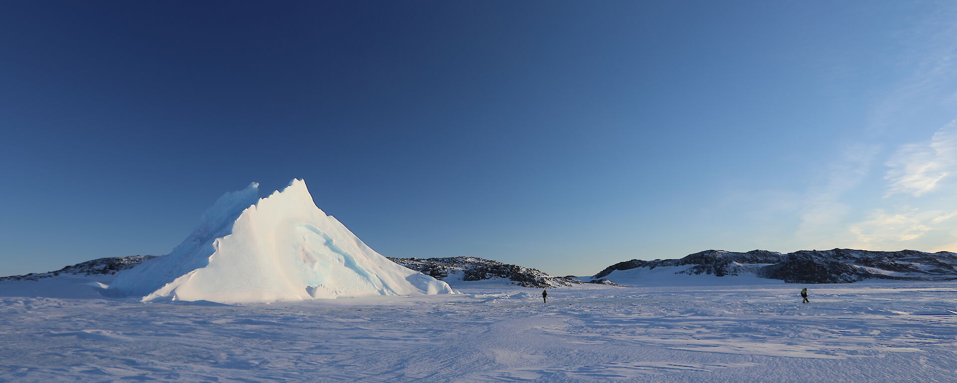 Expanse of sea-ice with pyramid ice berg sticking up out of it, two people walking across sea-ice towards the burg. Bright blue sky above, rocky hills in background